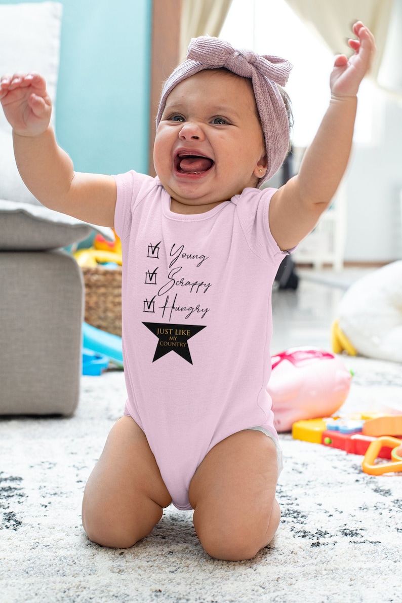 Pode incluir: Um macac&atilde;o rosa para beb&ecirc; com uma estrela preta e o texto "Young, Scrappy, Hungry, Just Like My Country".