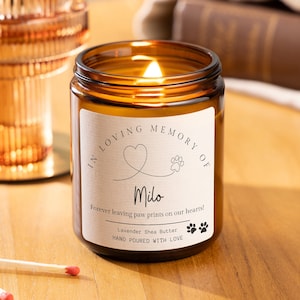 May include: A lit candle in an amber glass jar with a white label that reads In Loving Memory of Milo, Forever leaving paw prints on our hearts, Lavender Shea Butter, and Hand Poured With Love, featuring a heart and paw print graphic.