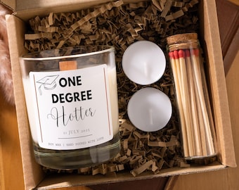 May include: A graduation gift box with a candle, two tea lights, and a matchstick tube. The candle has a label that reads "ONE DEGREE Hotter" with a graduation cap illustration and the date "21 JULY 2025". The box is filled with brown crinkle paper.