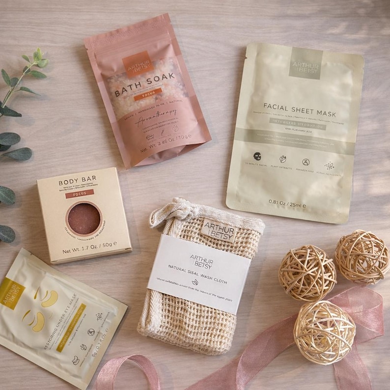 May include: A selection of self-care items, including a bath soak, facial sheet mask, body bar, and sisal wash cloth. The bath soak is 109g, and the body bar is 50g. The products are arranged with decorative woven spheres and a pink ribbon.
