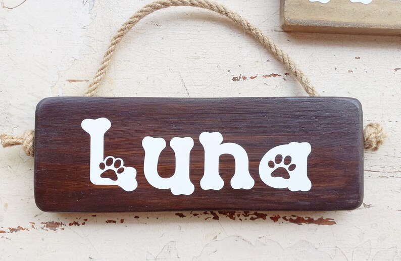 Pode incluir: Um sinal de madeira marrom escuro com um cabide de corda. O sinal tem o nome "Luna" escrito em branco com pegadas de patas de cada lado do nome.
