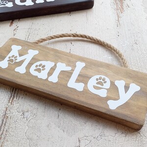Pode incluir: Um letreiro de madeira com o nome "Marley" escrito em letras brancas com pegadas de patas. O letreiro est&aacute; pendurado em uma corda e tem um visual r&uacute;stico.