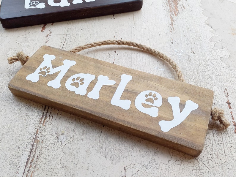 Pode incluir: Um letreiro de madeira com o nome "Marley" escrito em letras brancas com pegadas de patas. O letreiro est&aacute; pendurado em uma corda e tem um visual r&uacute;stico.