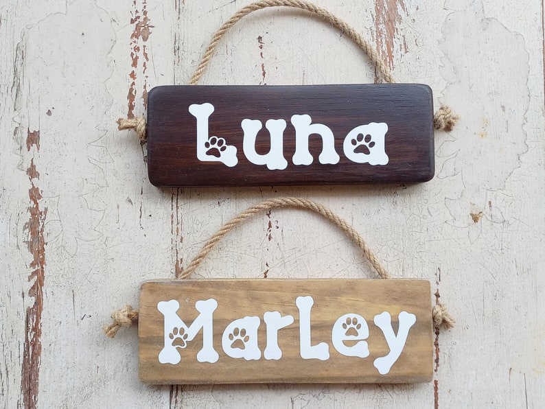 Pode incluir: Dois letreiros de madeira para c&atilde;es com pegadas de patas. O letreiro de cima &eacute; castanho escuro com o nome "Luna" e o letreiro de baixo &eacute; castanho claro com o nome "Marley".