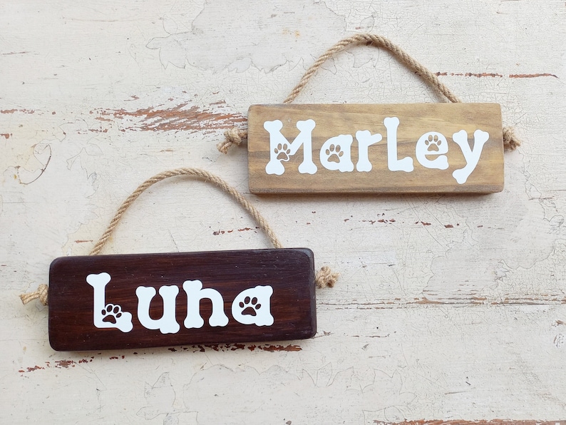 Pode incluir: Dois letreiros de madeira para c&atilde;es com pegadas de patas. O letreiro superior &eacute; de cor castanho claro com o nome "Marley" em branco. O letreiro inferior &eacute; de cor castanho escuro com o nome "Luna" em branco.
