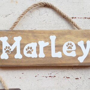 Pode incluir: Um sinal de madeira com o nome "Marley" escrito em letras brancas com pegadas de patas. O sinal est&aacute; pendurado em uma corda.