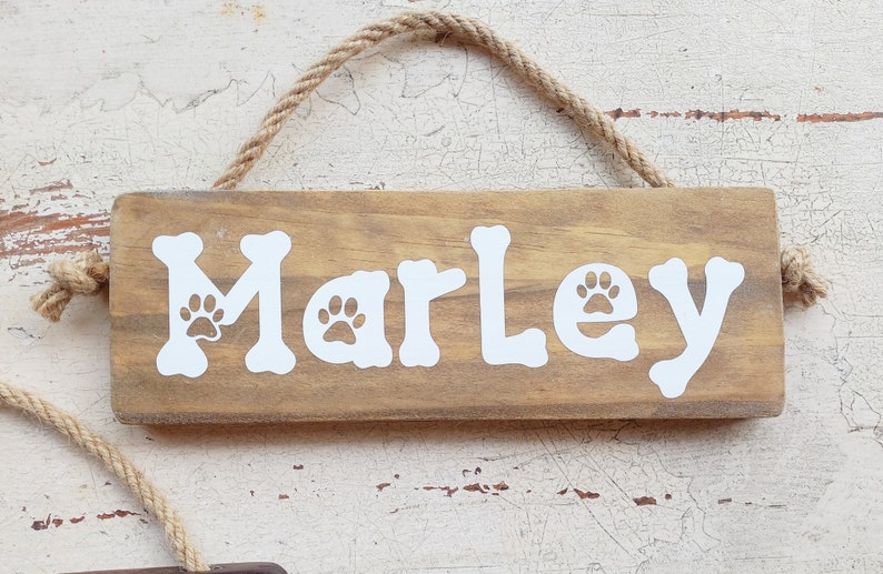 Pode incluir: Um sinal de madeira com o nome "Marley" escrito em letras brancas com pegadas de patas. O sinal est&aacute; pendurado em uma corda.