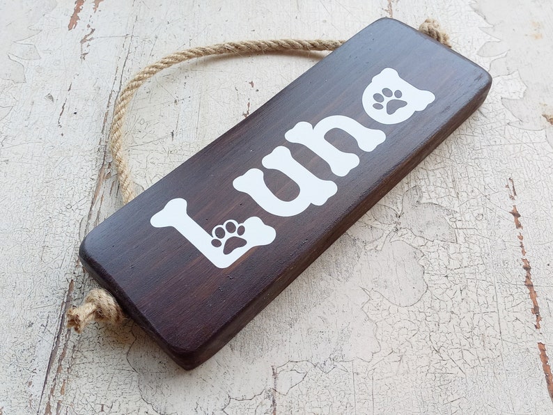 Pode incluir: Uma placa de madeira marrom escura com o nome "Luna" escrito em tinta branca. A placa tem duas pegadas brancas e est&aacute; pendurada em uma corda.