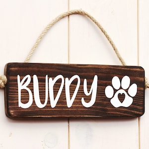 May include: A brown wooden sign with the word "BUDDY" and a white paw print with a heart in the center. The sign is hanging from a rope.