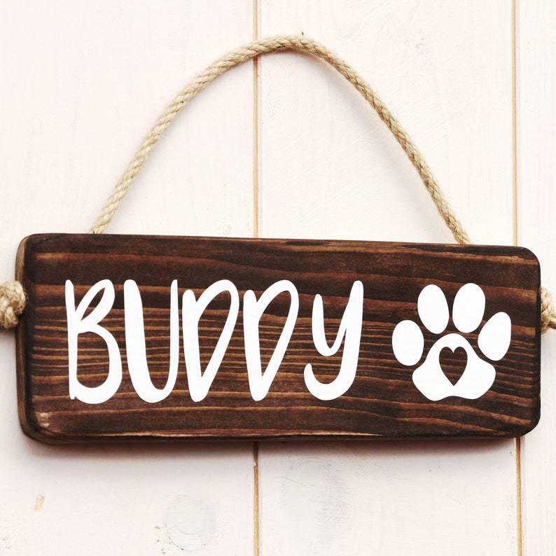 May include: A brown wooden sign with the word "BUDDY" and a white paw print with a heart in the center. The sign is hanging from a rope.