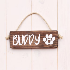 May include: A brown wooden sign with the word "BUDDY" and a white paw print with a heart inside. The sign is hanging from a piece of twine.