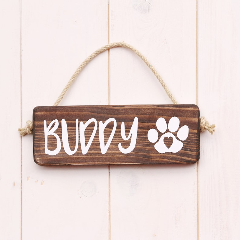 May include: A brown wooden sign with the word "BUDDY" and a white paw print with a heart inside. The sign is hanging from a piece of twine.