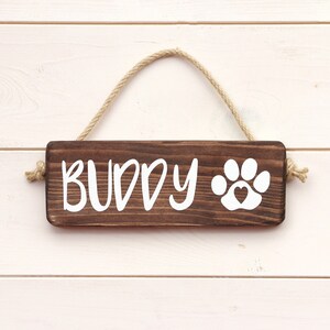 May include: A brown wooden sign with the word "BUDDY" and a white paw print with a heart inside. The sign is hanging from a rope.