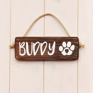 May include: A brown wooden sign with the word "BUDDY" and a white paw print with a heart in the center. The sign is hanging from a rope.