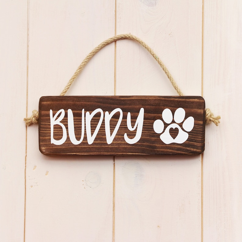 May include: A brown wooden sign with the word "BUDDY" and a white paw print with a heart in the center. The sign is hanging from a rope.