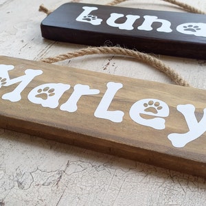Pode incluir: Dois letreiros de madeira com ganchos de corda, um marrom e outro de madeira natural. O letreiro marrom diz "Luna" em letras brancas com uma pegada de pata de cada lado. O letreiro de madeira natural diz "Marley" em letras brancas com uma pegada de pata de cada lado.