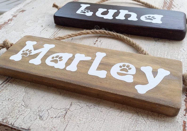 Pode incluir: Dois letreiros de madeira com ganchos de corda, um marrom e outro de madeira natural. O letreiro marrom diz "Luna" em letras brancas com uma pegada de pata de cada lado. O letreiro de madeira natural diz "Marley" em letras brancas com uma pegada de pata de cada lado.