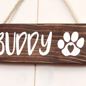 May include: A brown wooden sign with white text that reads "BUDDY" and a white paw print with a heart in the center. The sign is hanging from a rope.