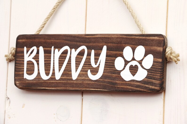 May include: A brown wooden sign with white text that reads "BUDDY" and a white paw print with a heart in the center. The sign is hanging from a rope.