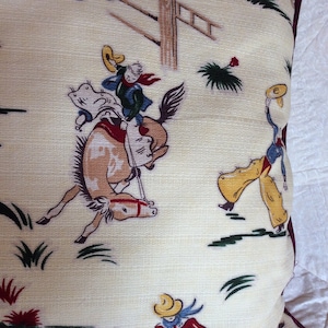 May include: A cream-colored pillowcase with a repeating pattern of cowboys riding horses. The cowboys are wearing colorful clothing and hats. The horses are brown and white. The background is a cream color with green grass and red flowers.