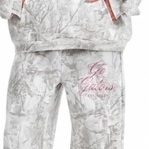May include: A matching two-piece set consisting of a hooded sweatshirt and wide-leg pants in a light gray and white camouflage leaf pattern. The set features the pink script text Go Gators and the text EST. 1853 printed on the chest and left thigh.