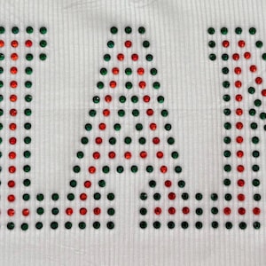 K&ouml;nnte beinhalten: Wei&szlig;er Stoff mit dem Wort "MIAMI" aus roten und gr&uuml;nen Strasssteinen. Die Buchstaben bestehen aus zahlreichen kleinen, funkelnden Strasssteinen. Der Stoff hat eine subtile Rippenstruktur, wobei das Wort deutlich sichtbar ist.