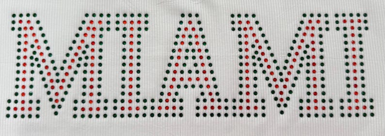 K&ouml;nnte beinhalten: Wei&szlig;er Stoff mit dem Wort "MIAMI" aus roten und gr&uuml;nen Strasssteinen. Die Buchstaben bestehen aus zahlreichen kleinen, funkelnden Strasssteinen. Der Stoff hat eine subtile Rippenstruktur, wobei das Wort deutlich sichtbar ist.