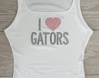May include: White ribbed tank top with a square neckline. The front features the phrase "I ❤️ GATORS" in black and red rhinestones. The top is made of a soft, ribbed material.