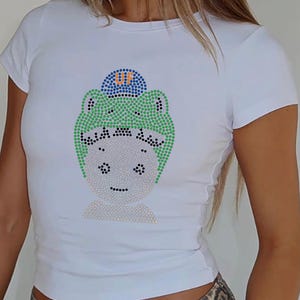 Puede incluir: Camiseta blanca con un dise&ntilde;o de pedrer&iacute;a de un personaje de dibujos animados que lleva un gorro verde y un gorro azul con las letras "UF". El dise&ntilde;o est&aacute; hecho de pedrer&iacute;a de varios colores. La camiseta tiene mangas cortas.