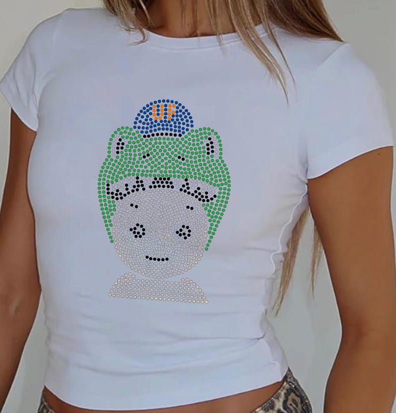 Puede incluir: Camiseta blanca con un dise&ntilde;o de pedrer&iacute;a de un personaje de dibujos animados que lleva un gorro verde y un gorro azul con las letras "UF". El dise&ntilde;o est&aacute; hecho de pedrer&iacute;a de varios colores. La camiseta tiene mangas cortas.