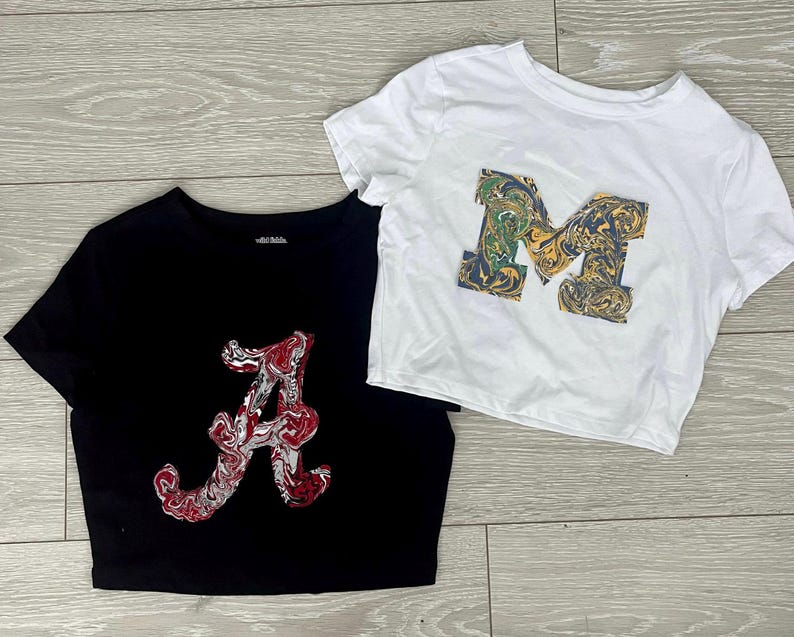 Puede incluir: Dos crop tops negros y blancos con una letra impresa en la parte delantera. El top de la izquierda tiene una letra "A" roja y blanca y el top de la derecha tiene una letra "M" verde, amarilla y blanca.