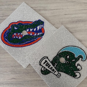 Könnte beinhalten: Zwei silberne Strass-Untersetzer. Einer zeigt einen grünen und blauen Alligatorkopf mit orangefarbenem und blauem Umriss. Der andere hat ein grünes Wesen mit einer blauen Welle und einer Trompete mit der Aufschrift "TULANE".