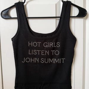 May include: Black ribbed tank top with the text "HOT GIRLS LISTEN TO JOHN SUMMIT" in silver rhinestones. The top is hanging on a black hanger against a white door.