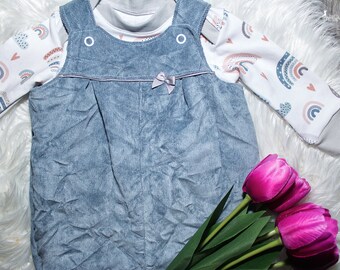 Op de afbeelding: Baby-outfitset met een blauwe corduroy overall met een kleine strik en een wit shirt met lange mouwen. Het shirt heeft een regenboog- en hartpatroon. De outfit wordt gepresenteerd op een witte, pluizige ondergrond, met roze tulpen op de voorgrond.