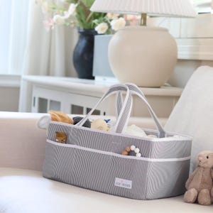 May include: A rectangular grey and white pinstriped storage caddy with two fabric handles and multiple exterior pockets. The front features a white label with the text Lily Miles. The interior contains various small items and toys.