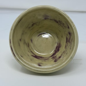 May include: A round ceramic bowl with a glossy finish, featuring a pale cream base color accented by swirling, irregular streaks of deep purple pigment throughout the interior surface and along the rounded rim of the vessel.
