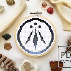 以下が含まれることがあります： 円の中にシンボルが描かれた白黒のクロスステッチパターン。シンボルは、上に3つの点がある三角形のスタイリッシュな画像です。パターンは木製の刺繍枠に入っています。