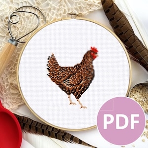 May include: A brown and white chicken cross stitch pattern in a wooden embroidery hoop. The pattern is on white Aida cloth and the chicken is brown with a red comb. The hoop is on a white lace tablecloth with a red spoon and a wooden rolling pin.