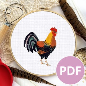 May include: A cross-stitch pattern of a colourful rooster with a red comb and wattle, a yellow body, a black and green tail, and a white background. The pattern is displayed in a round embroidery hoop.