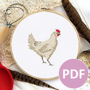 May include: A white chicken cross stitch pattern in a wooden embroidery hoop. The pattern is on white Aida cloth and features a white chicken with a red comb and wattle. The pattern is surrounded by a white lace doily and a wooden rolling pin.
