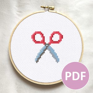 May include: Cross-stitch embroidery of red scissors with blue blades, set in a wooden hoop. The design is on white fabric. A pink circle with the letters "PDF" is in the lower right corner.