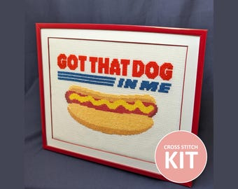 May include: Cross-stitch kit featuring a framed design with the text "GOT THAT DOG IN ME" above a pixelated hot dog with mustard. The kit includes a red frame and a pink circle with the words "CROSS STITCH KIT".