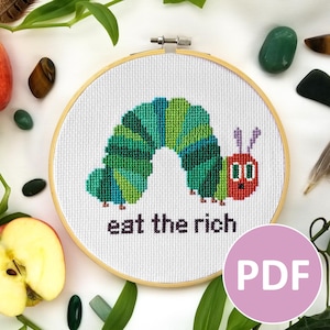 Könnte beinhalten: Ein Kreuzstich-Stickrahmen mit einer grün-blauen Raupe mit rotem Gesicht. Der Text "eat the rich" ist unter der Raupe gestickt. Das Bild ist von Äpfeln, Blättern und dekorativen Steinen umgeben.