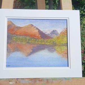 Peut inclure: Peinture de paysage à l'aquarelle de montagnes et d'un lac, encadrée en blanc. Les montagnes sont dans des tons orange et marron, avec un reflet dans l'eau bleue. Des arbres verts et jaunes bordent la rive. Le ciel est bleu clair.