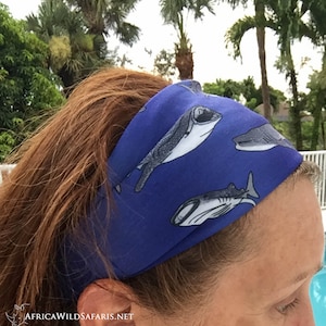 May include: A blue headband featuring a pattern of gray and white whale shark illustrations. The headband is worn on the head, with the hair visible. The background shows green trees and a white railing.