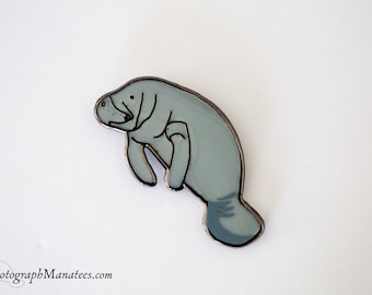 May include: A light blue manatee pin with a black outline. The manatee is in a swimming pose. The pin is made of enamel and has a shiny finish. A charming accessory, the pin depicts a manatee, a marine mammal, in a whimsical style.