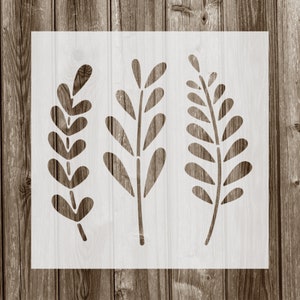May include: A white stencil featuring three botanical designs of leaves and branches. The stencil is rectangular and sits on a wooden surface with vertical planks. The leaves have rounded and elongated shapes, perfect for DIY projects.