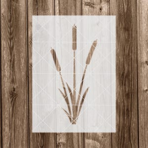 May include: A stencil of three brown cattail plants with long, thin leaves. The stencil is on a white background.
