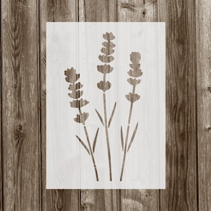 May include: A white stencil with a design of three lavender flowers. The stencil is on a brown wooden background.