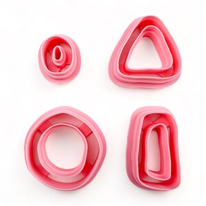 May include: Four pink plastic cookie cutters in different shapes: a circle, a rectangle, a triangle, and a smaller circle. The cutters are designed for making cookies or other pastries.